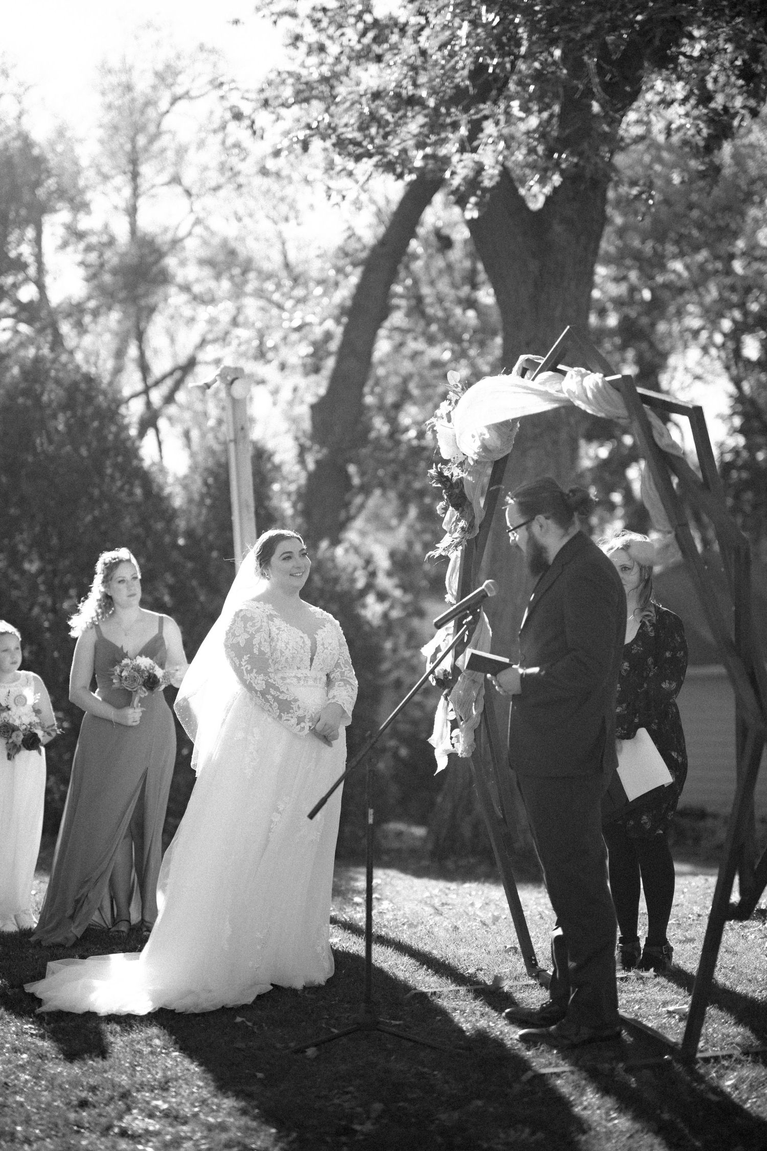 Beautiful outdoor wedding ceremony in a sunlit garden with wedding party standing beneath trees in black and white photography.