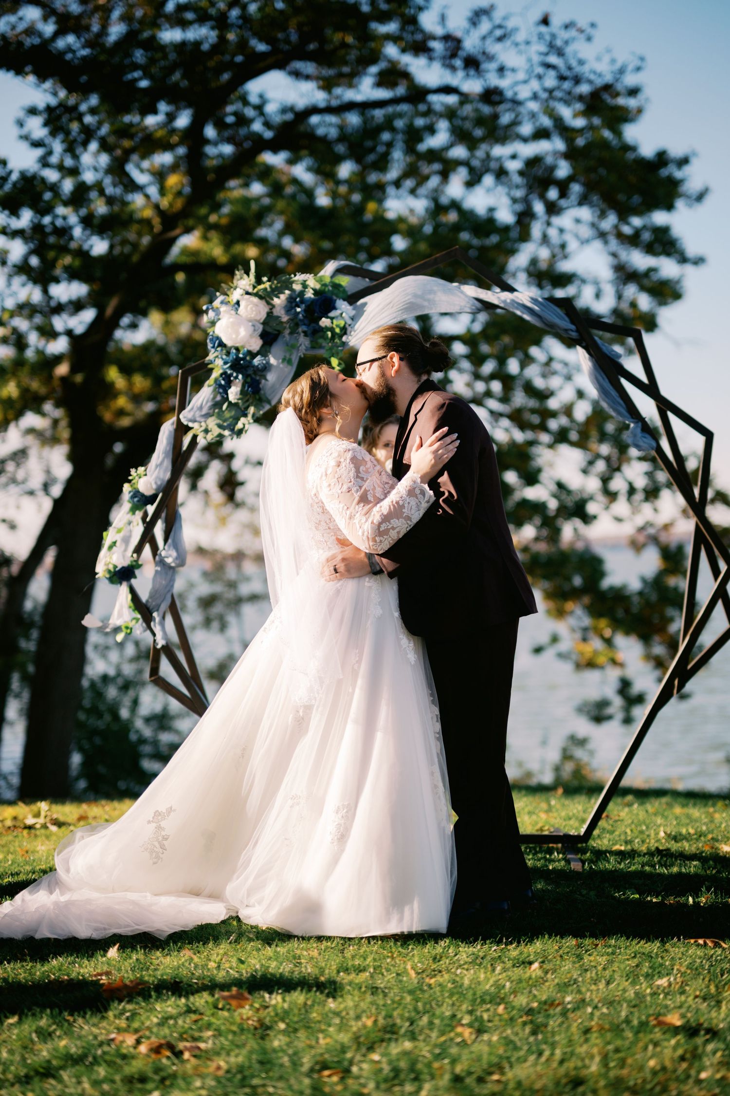 A romantic outdoor wedding ceremony with a couple kissing under a floral arch decorated with white flowers on a grassy hilltop.