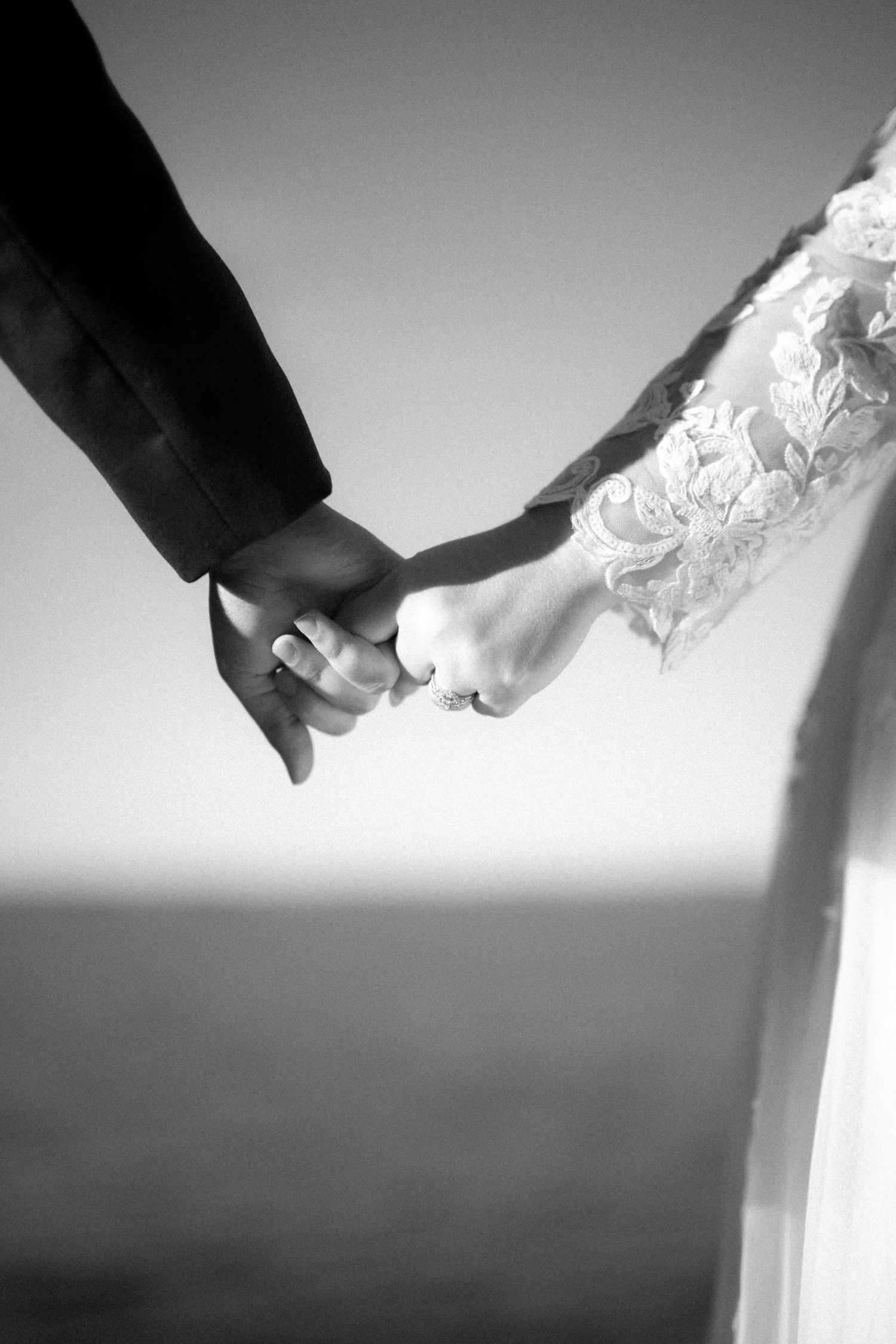 Black and white closeup of newlyweds holding hands, with the bride wearing delicate lace sleeves and the groom in dark attire.