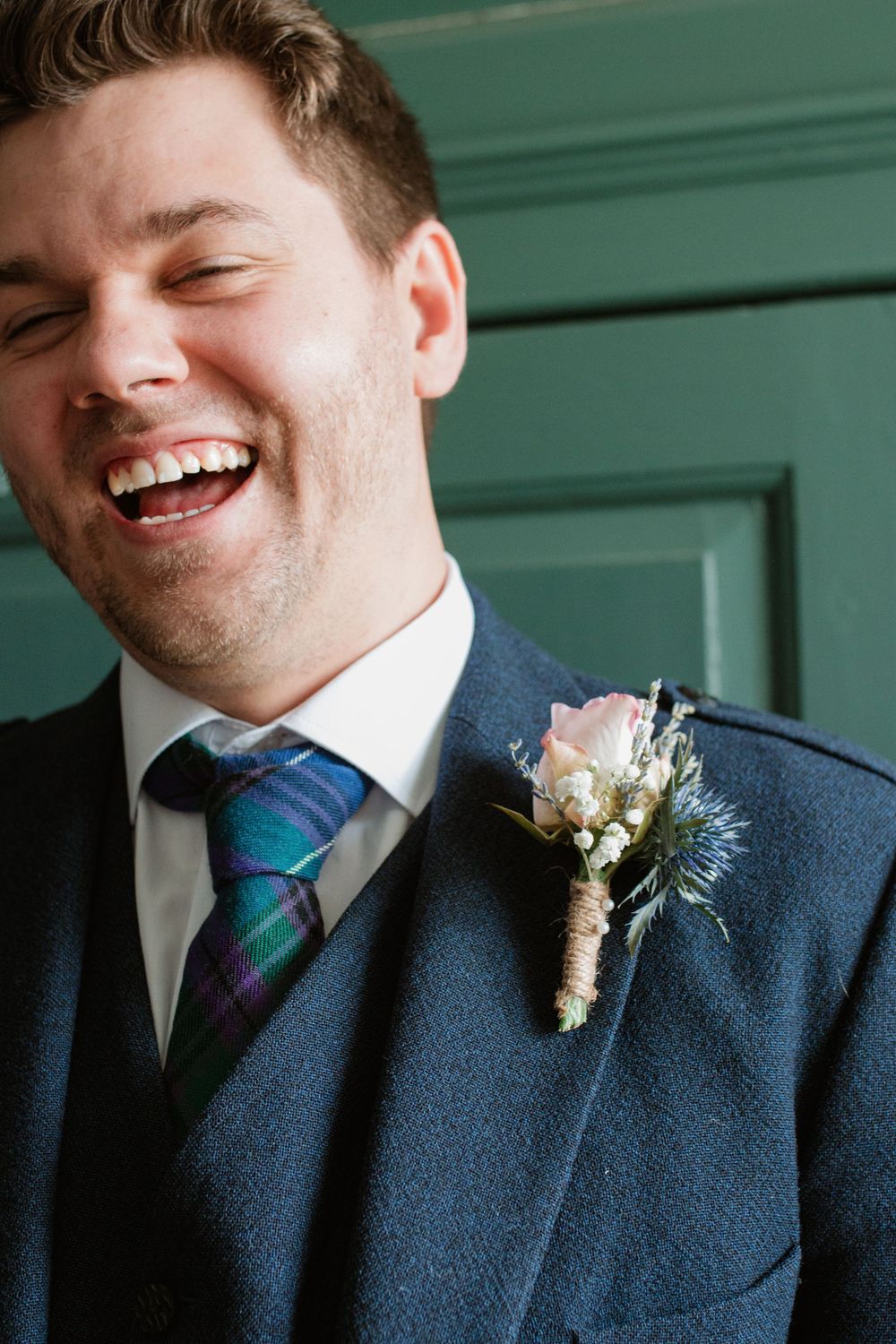 Groom in navy suit with plaid tie and boutonniere smiles against green wall.
