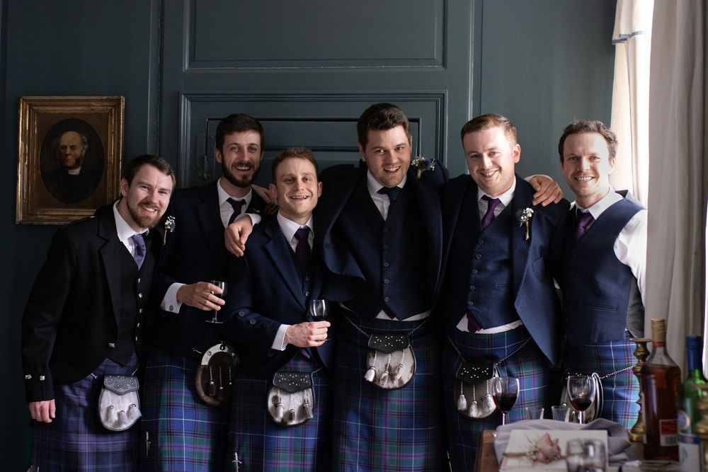 Groomsmen in matching navy suits stand together holding drinks.