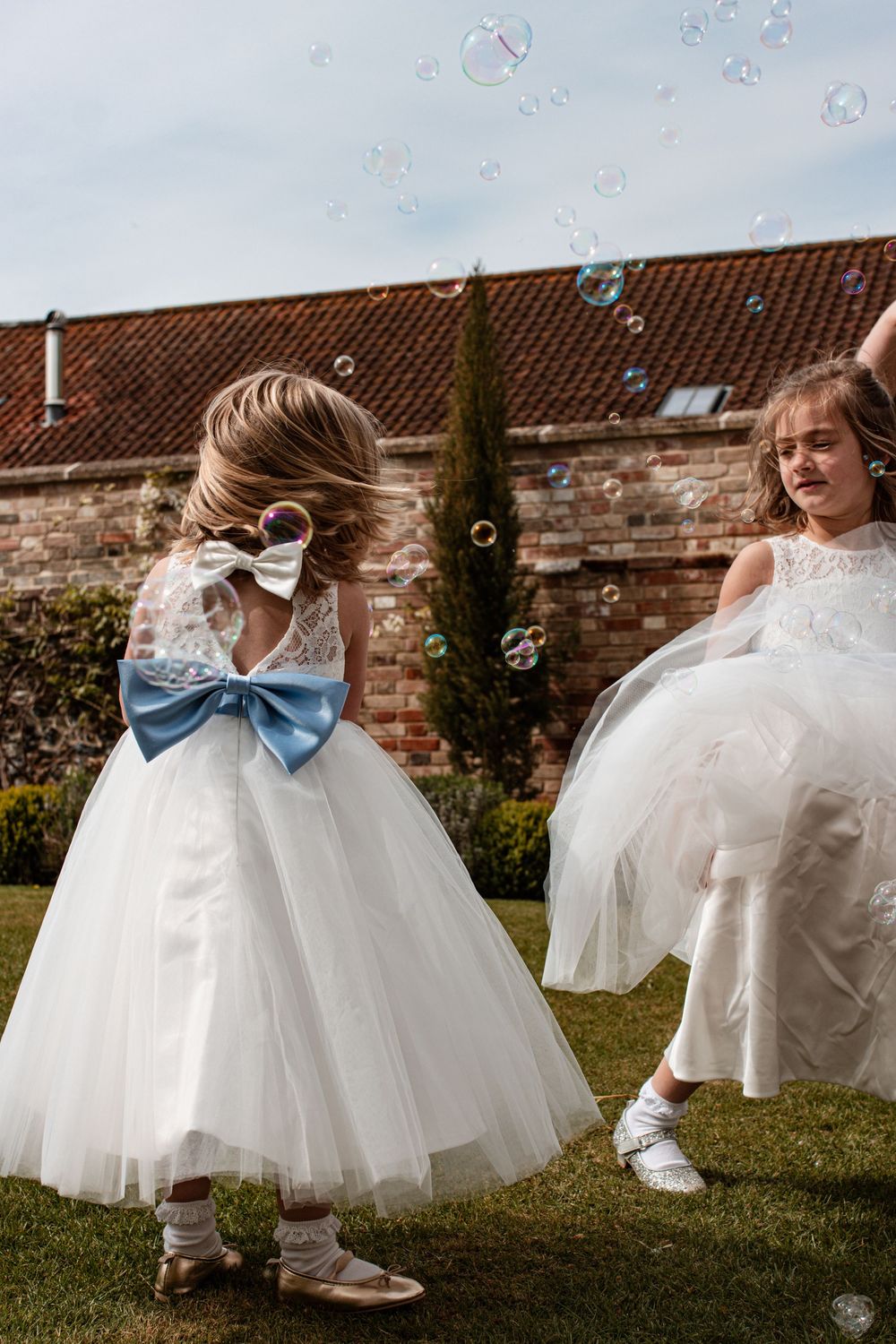 Young attendants in white dresses with blue bows play with bubbles on a grassy lawn.