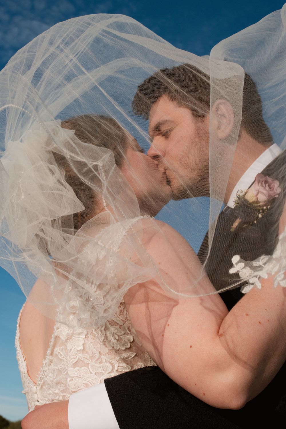 A dreamy wedding kiss captured against a bright blue sky with flowing veil details.