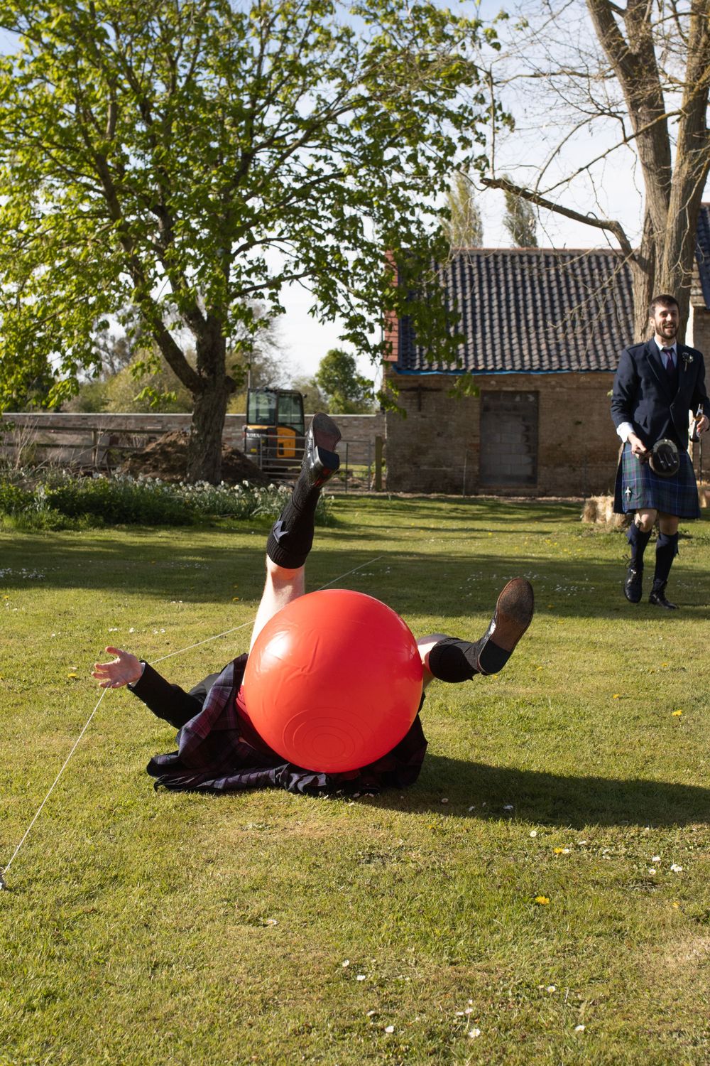 Someone in formal attire rolls playfully on a large red balloon in the grass.