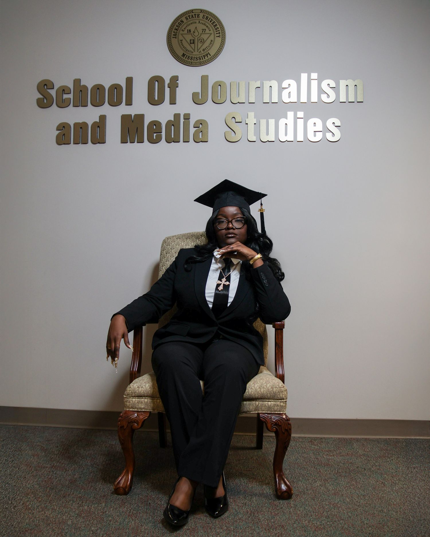 Graduate in cap and gown poses in front of School of Journalism and Media Studies sign.
