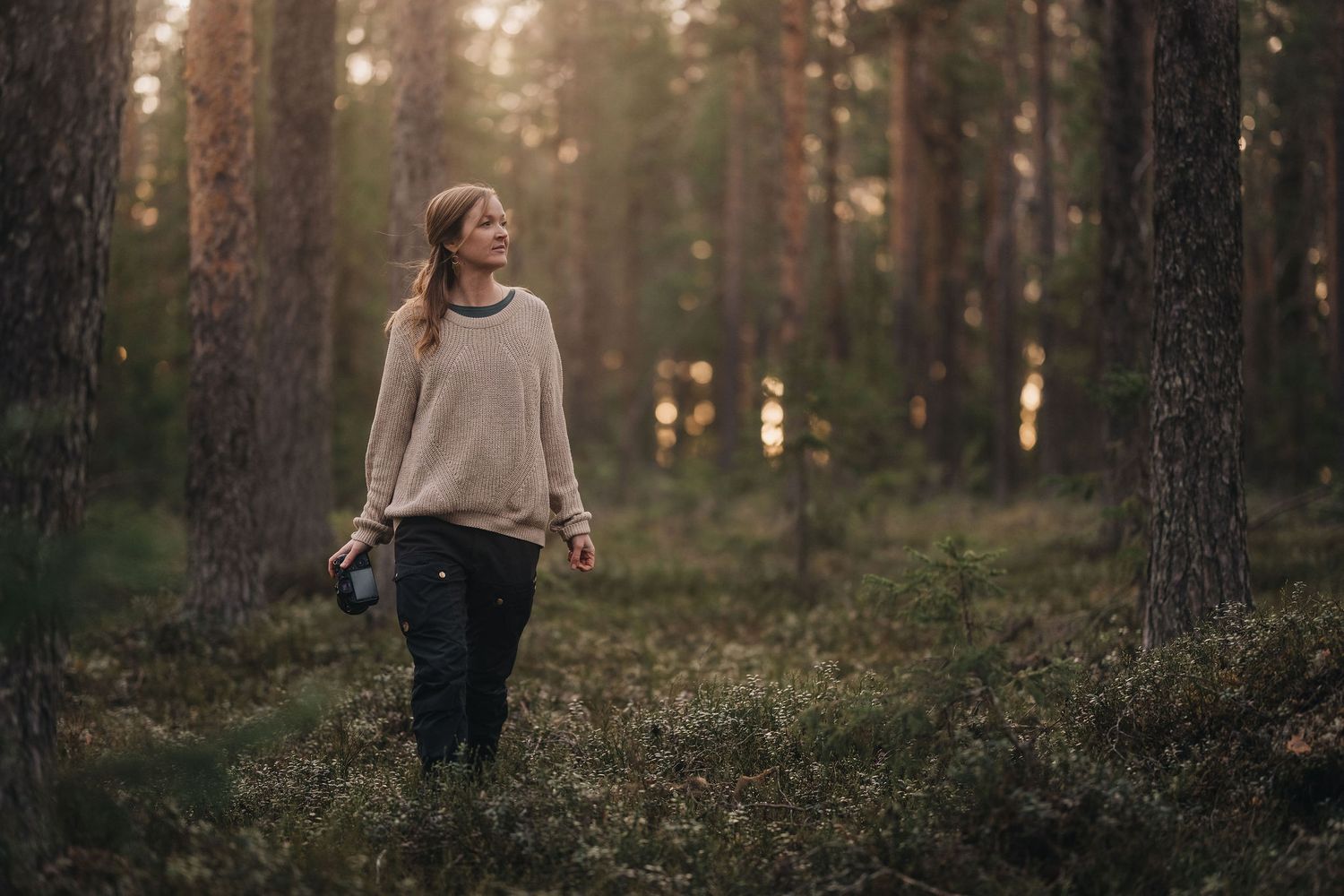 Woman in beige sweater walking through pine forest at dusk holding camera.