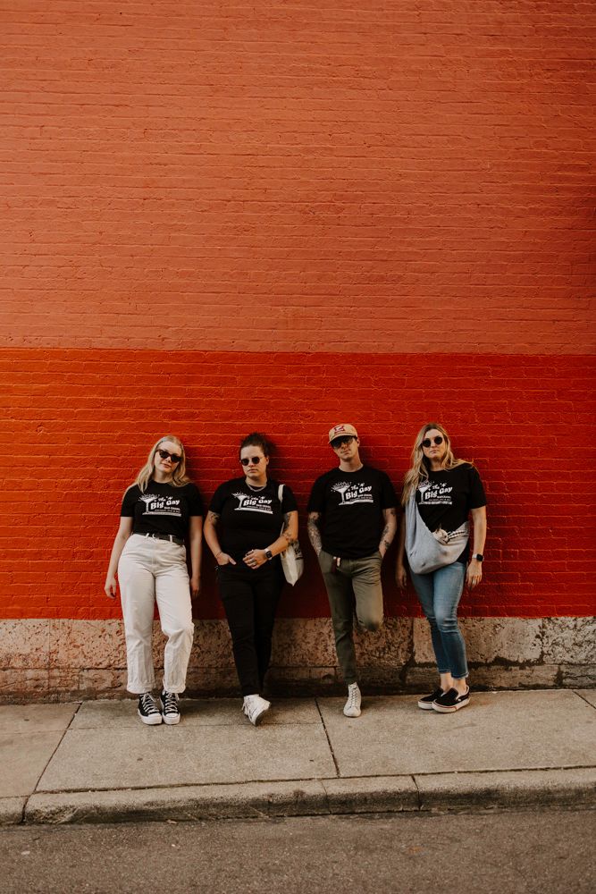 Four people in matching black t-shirts standing against an orange painted wall.