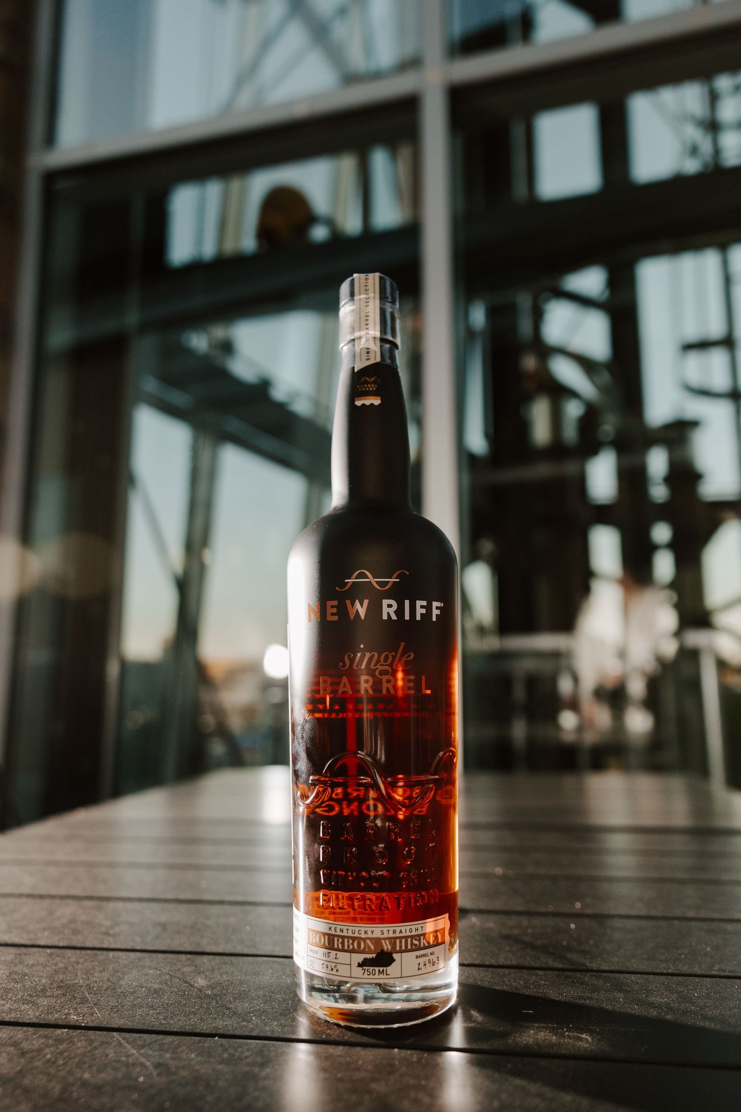 Bottle of New Riff bourbon whiskey displayed on wooden table with industrial windows in background.