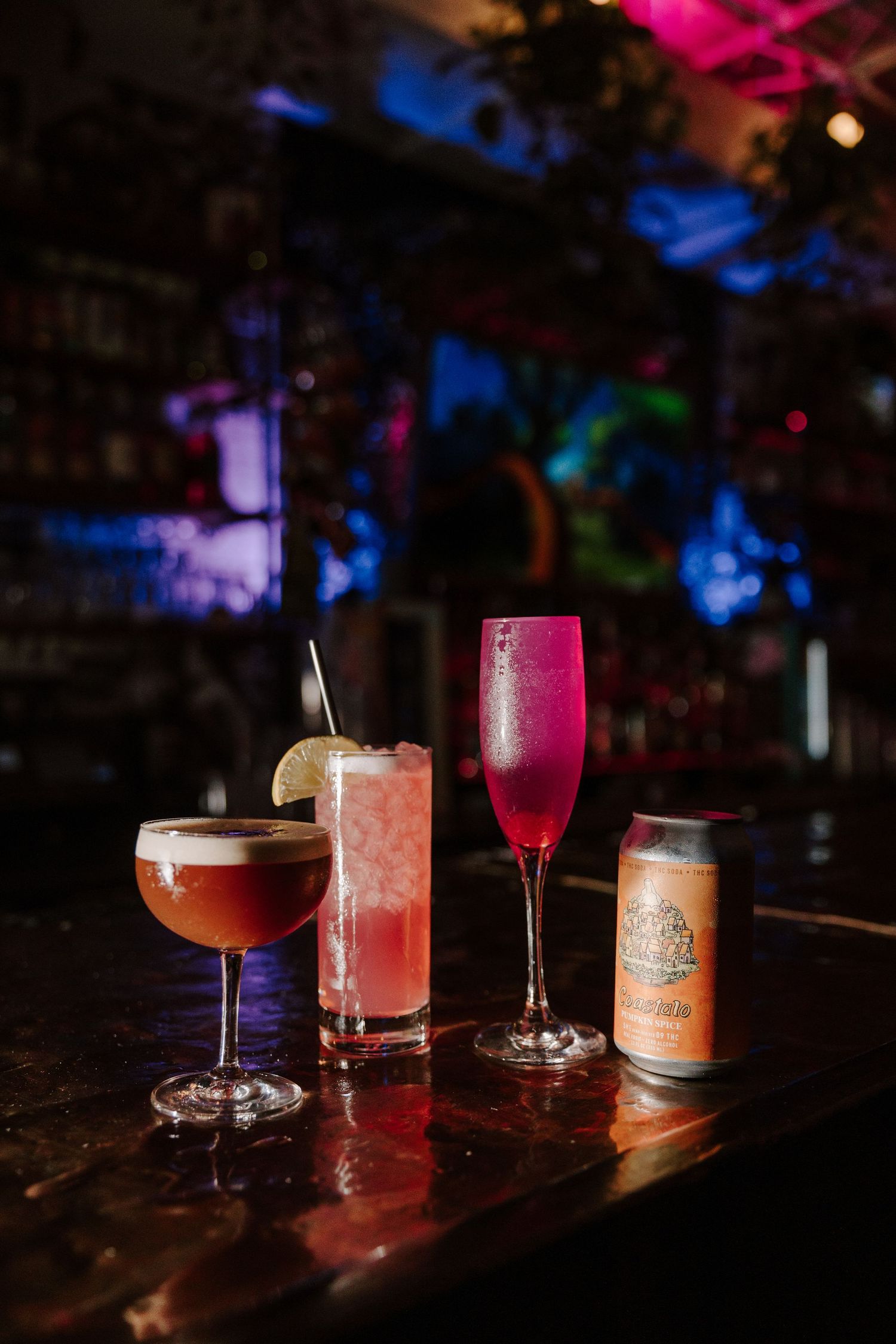 Assorted cocktails and beverages displayed on dark bar top with colorful ambient lighting.