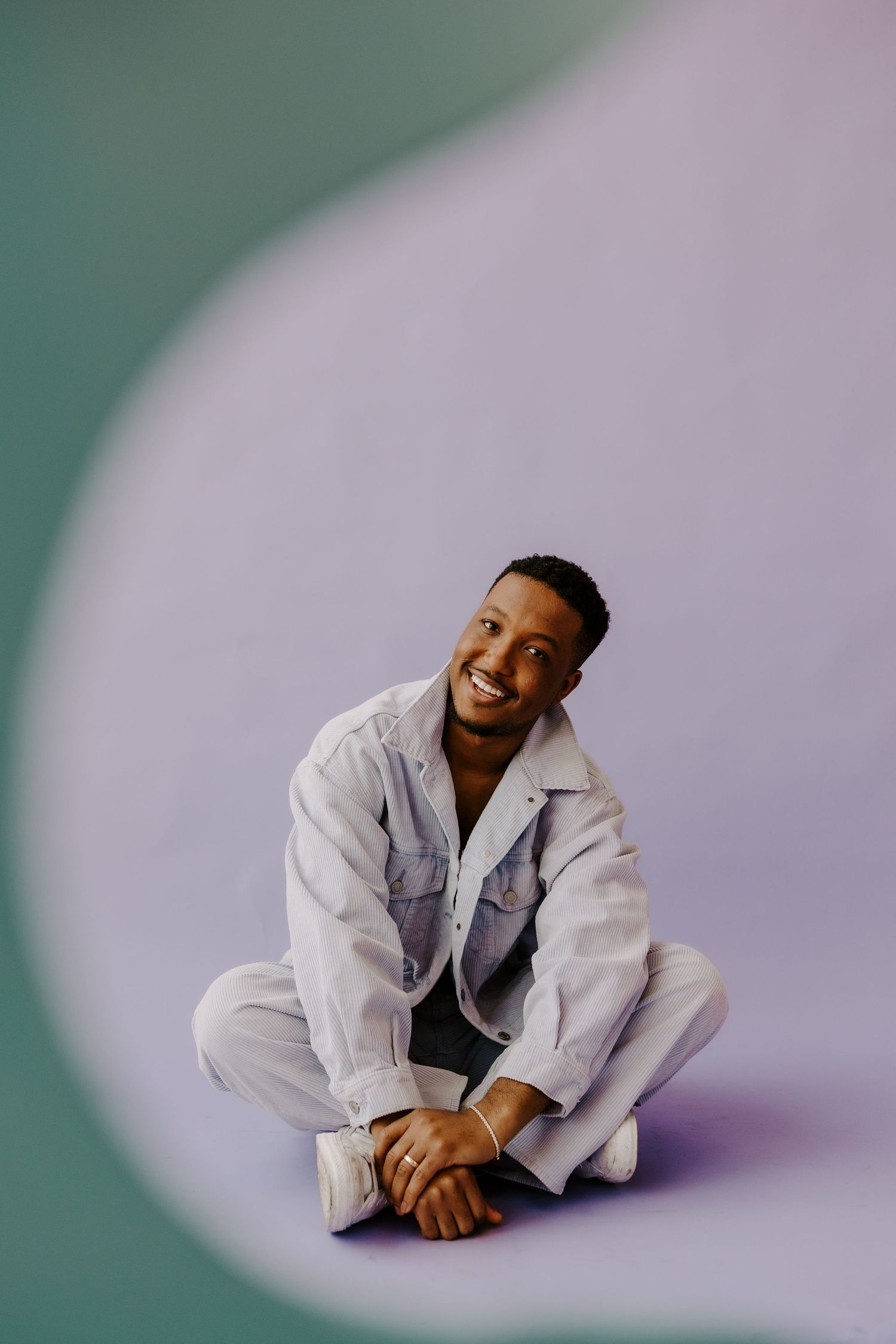 Person in white denim outfit sits cross-legged and smiles warmly in a minimalist studio setting.