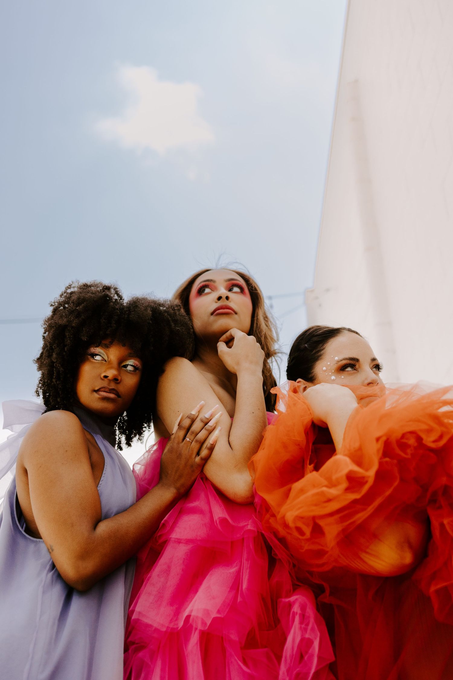 Three friends in vibrant dresses of purple, pink and orange pose artistically against a light sky background.