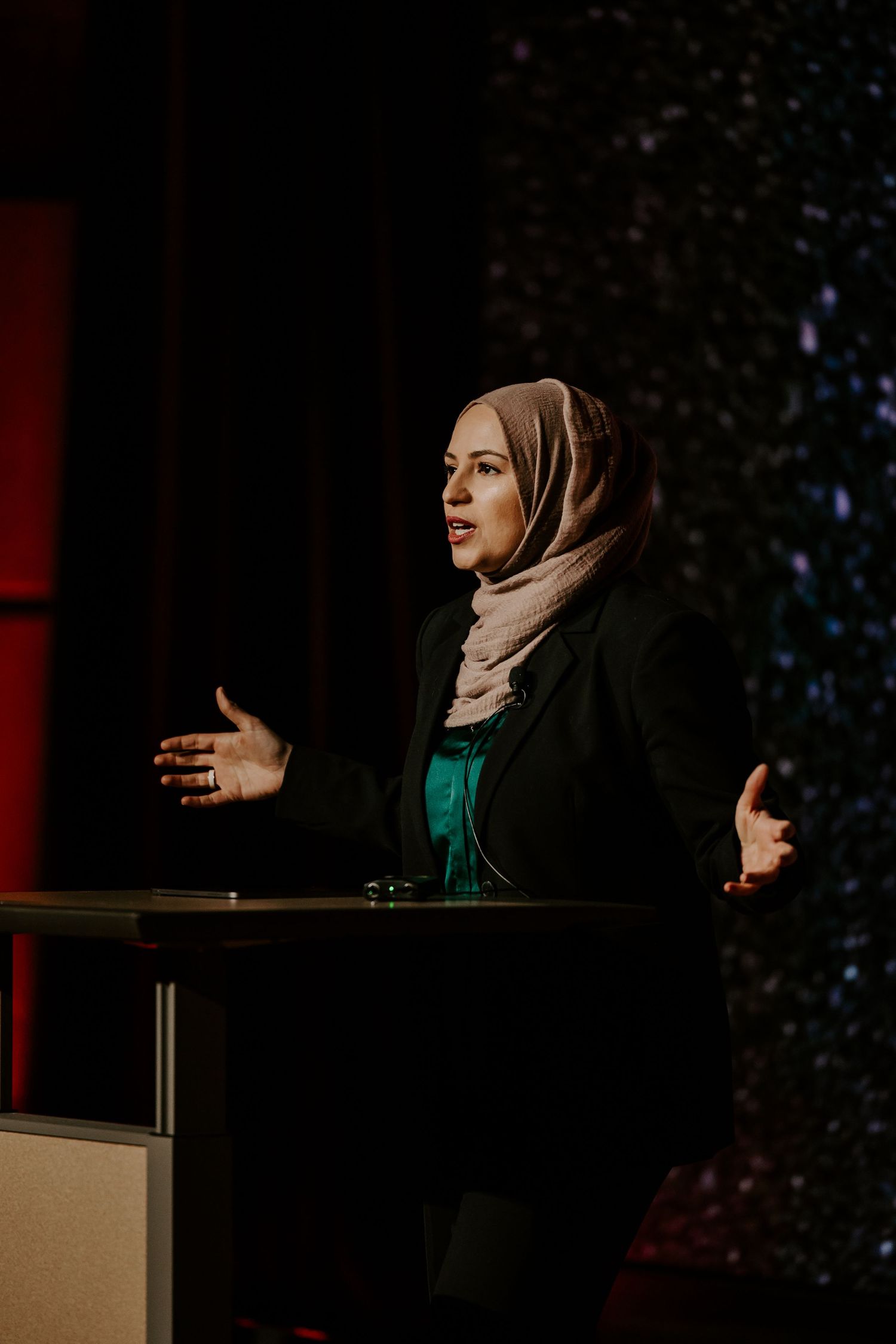 Speaker in hijab and professional attire gives presentation at podium with dramatic lighting.