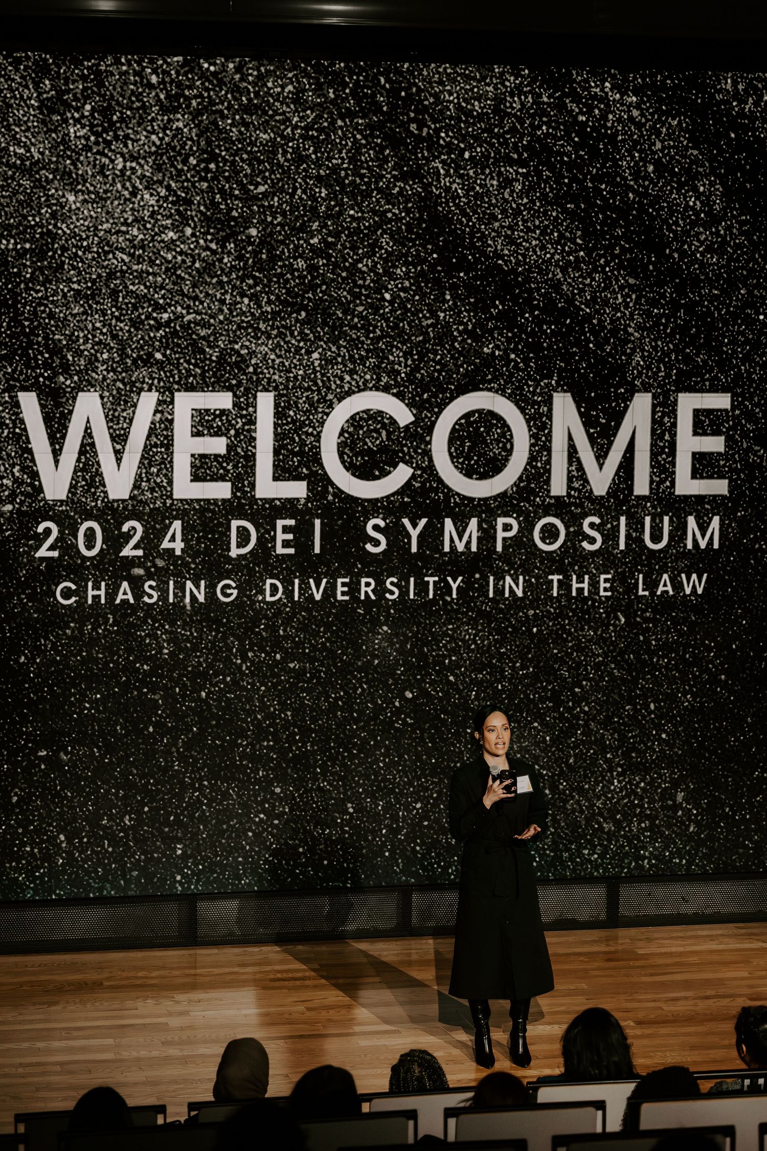 Speaker at diversity in law symposium standing beneath welcome sign on dark sparkly background.