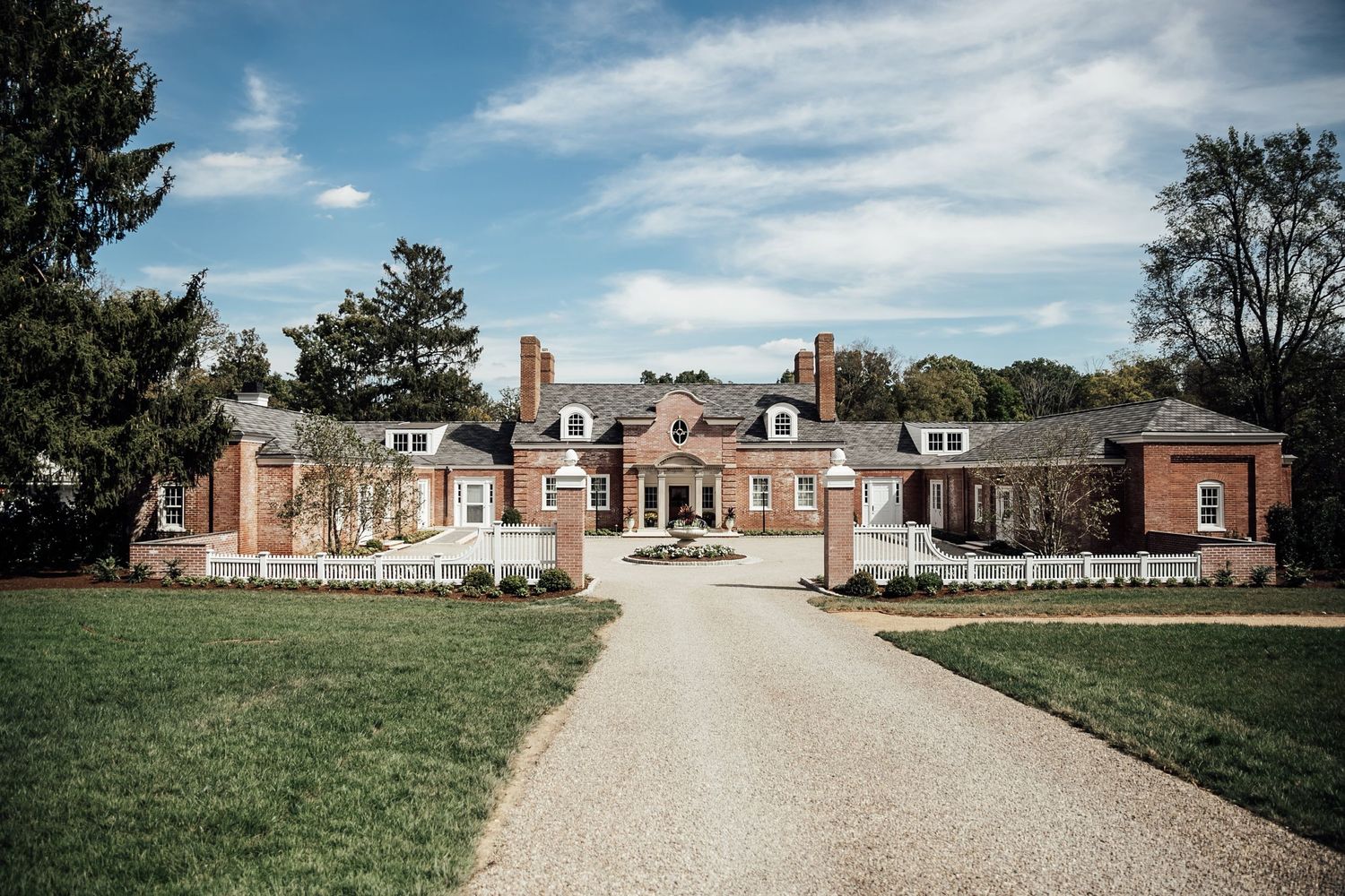 Grand brick mansion with colonial architecture and gravel driveway under blue skies.