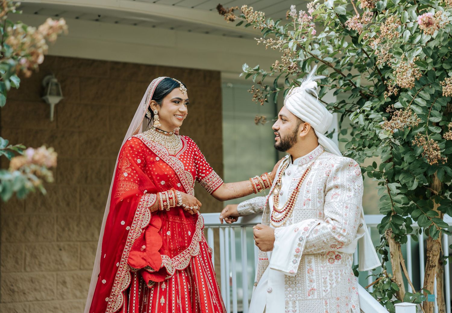 Indian bride in red embroidered lehenga and groom in cream sherwani with turban smile at each other during a wedding portrait outdoors.