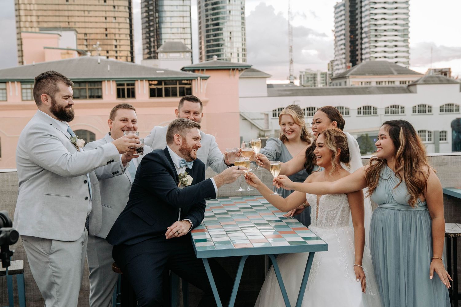 Wedding party toasts with drinks on a rooftop venue with city buildings in background.