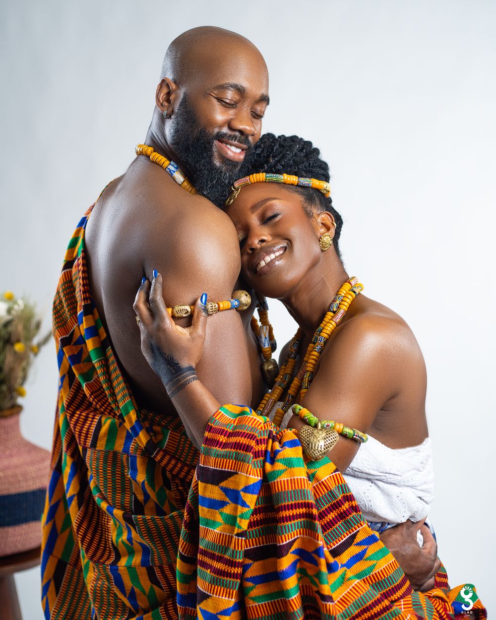 A loving couple dressed in vibrant kente cloth embraces tenderly against a light background.