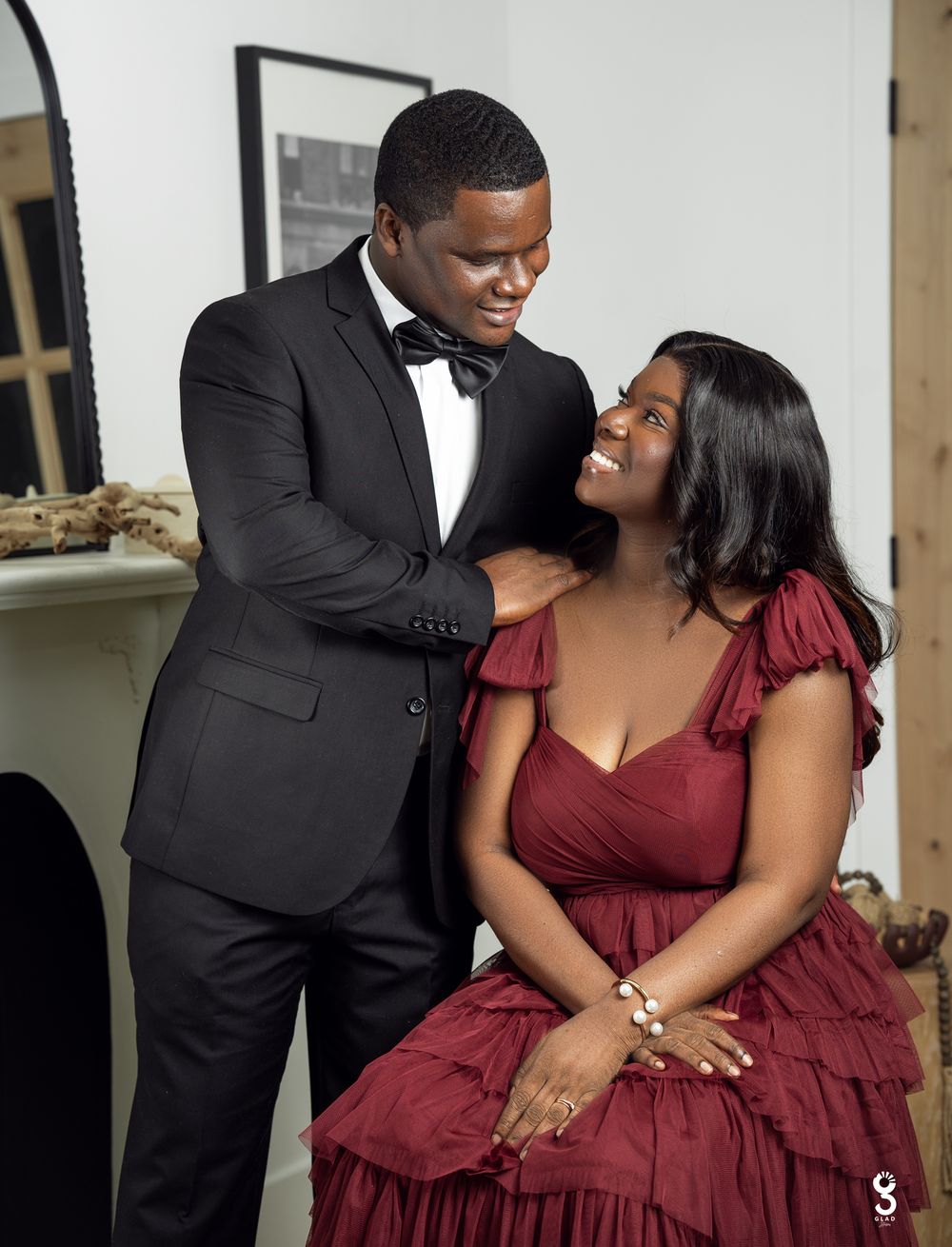 A couple shares a tender moment, with the person in a black tuxedo looking lovingly at their partner in a burgundy gown.