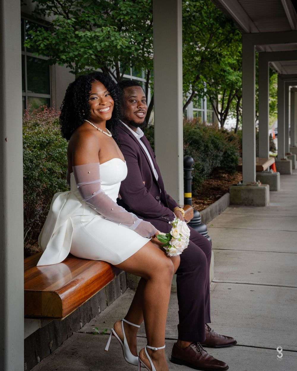 A couple in formal attire poses together in an outdoor covered walkway with columns and wooden benches.