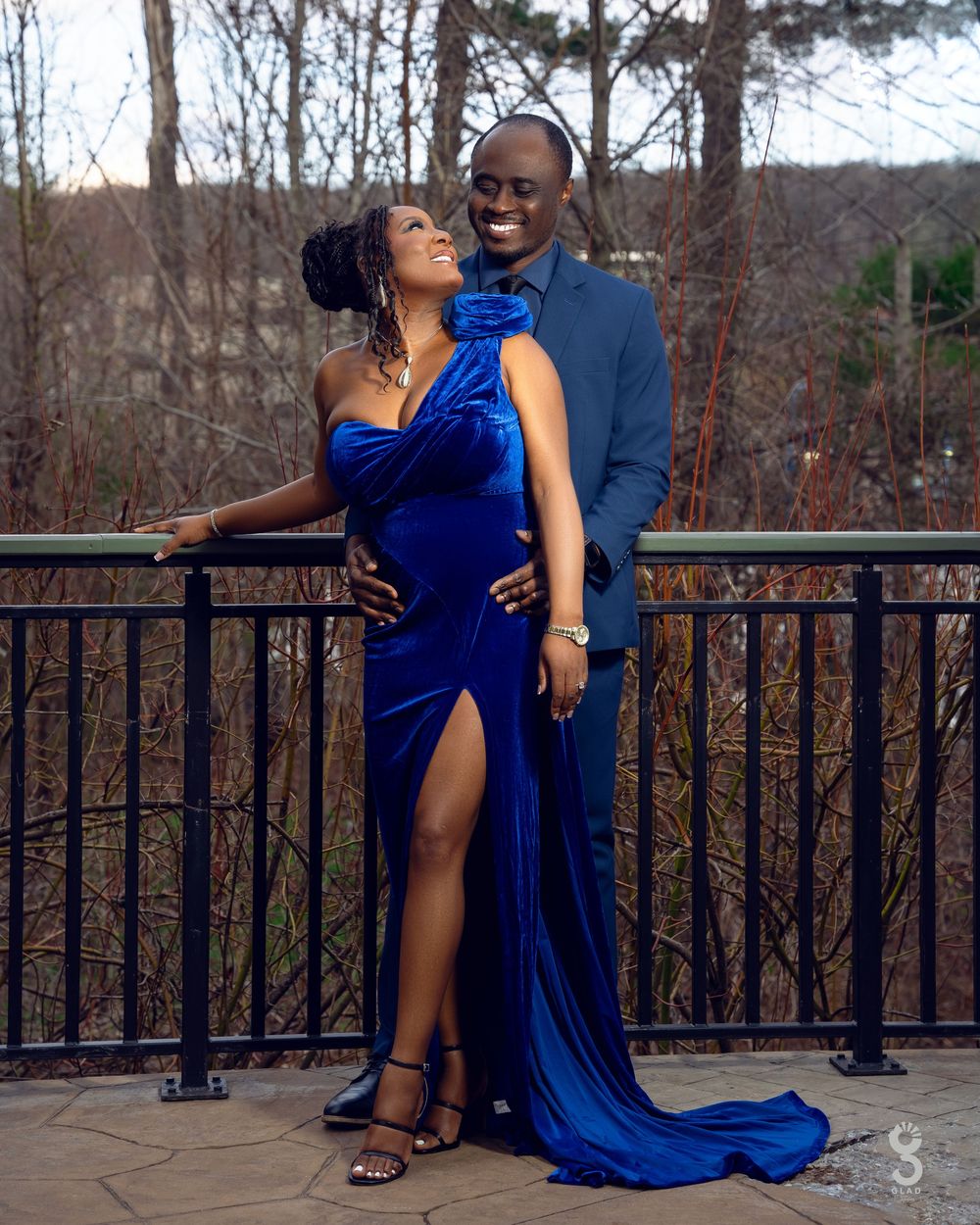 A couple in formal attire poses on a deck with a scenic wooded background as the blue dress flows elegantly.