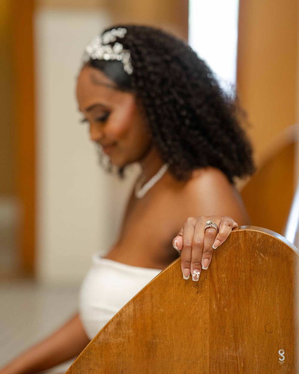A bride in a white dress with natural curly hair and a delicate hairpiece rests her hand on a wooden chair.