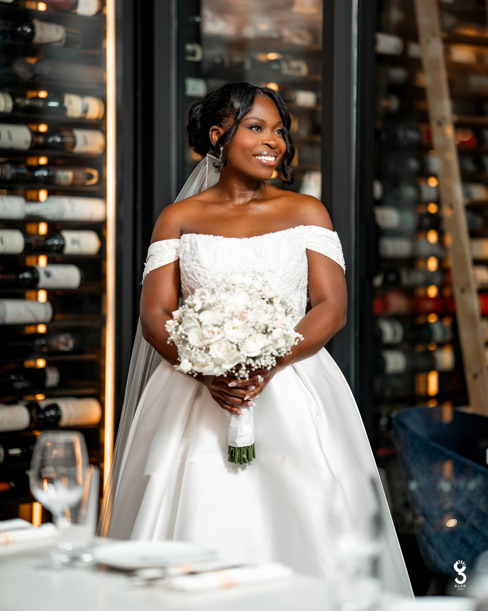 Beautiful bride in off-shoulder white gown holds white bouquet while standing in front of wine cellar display.