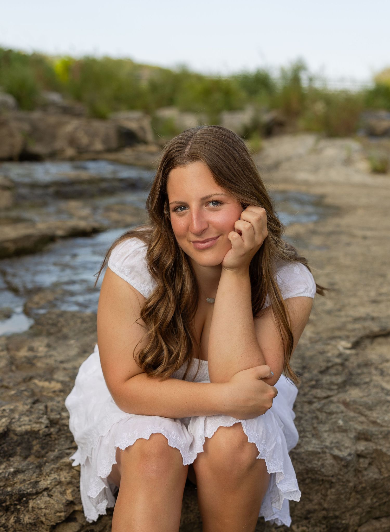 Natural portraits of woman in white dress sitting on rocky shoreline.