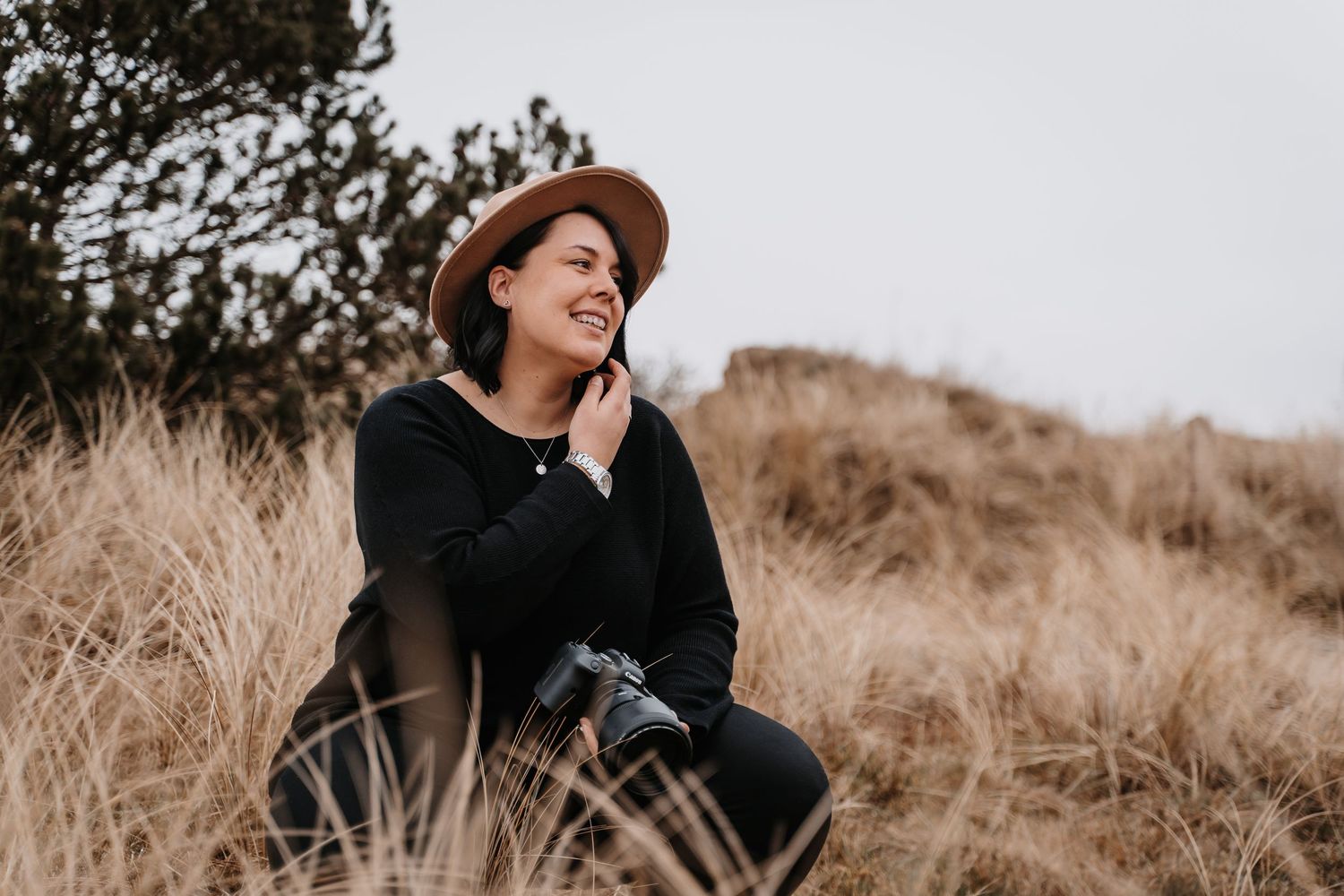 Woman in black sweater and brown hat sitting in tall autumn grass.