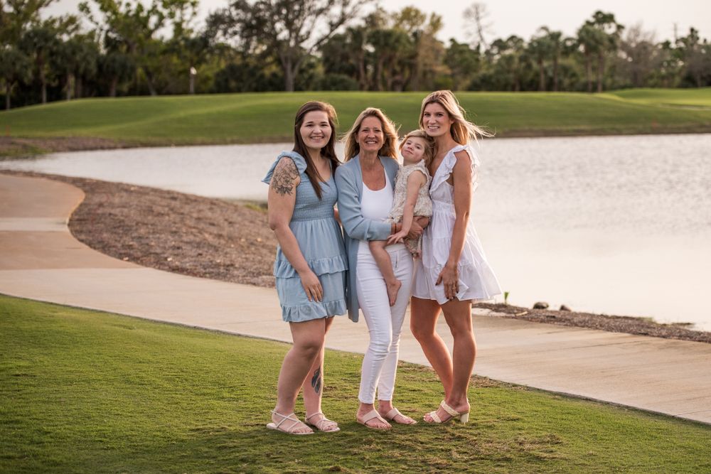 Three women in light summer dresses pose together by a lake during golden hour.