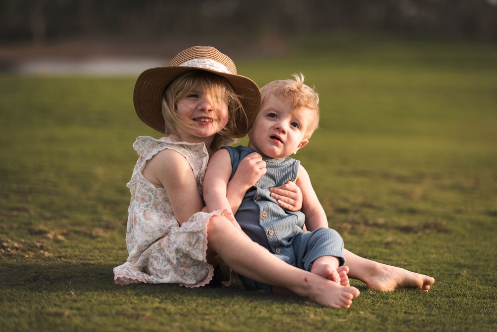 Two children relax outdoors on grass with one wearing a straw sunhat during golden hour.