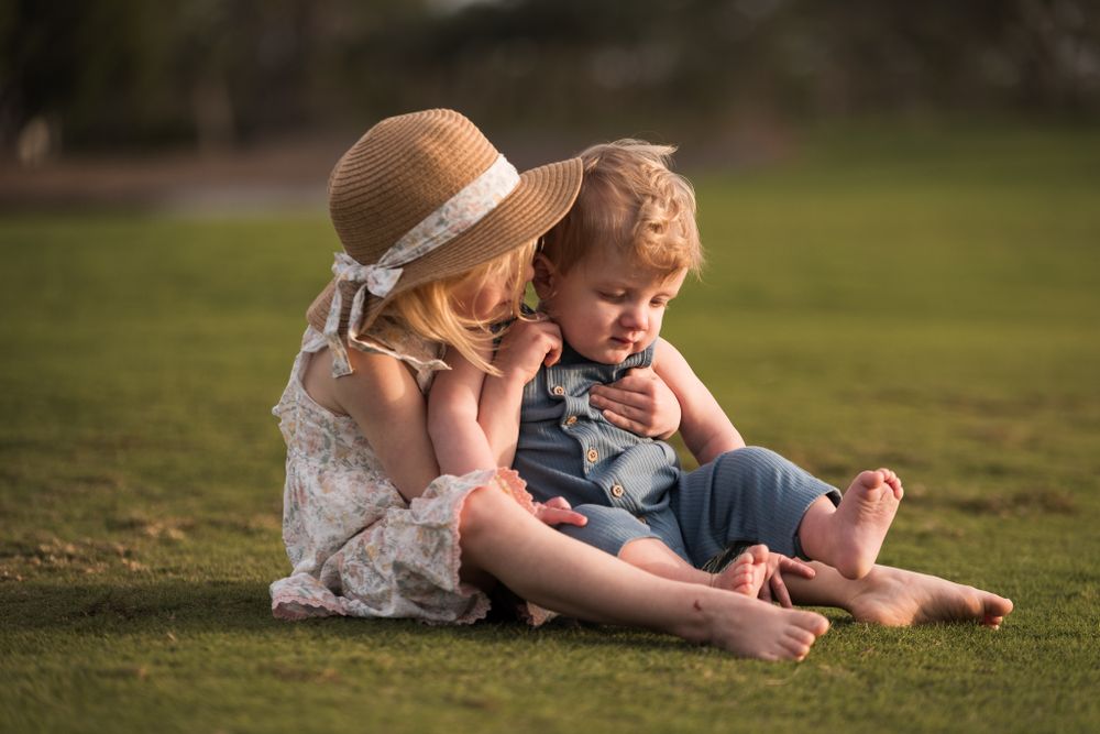 Two children sit on grass during sunset with one wearing a straw hat and white dress.