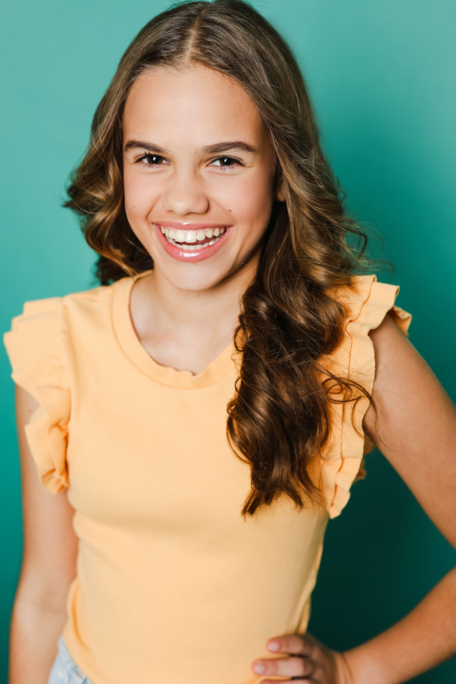Young person in yellow ruffled top smiling against turquoise background in professional headshot.