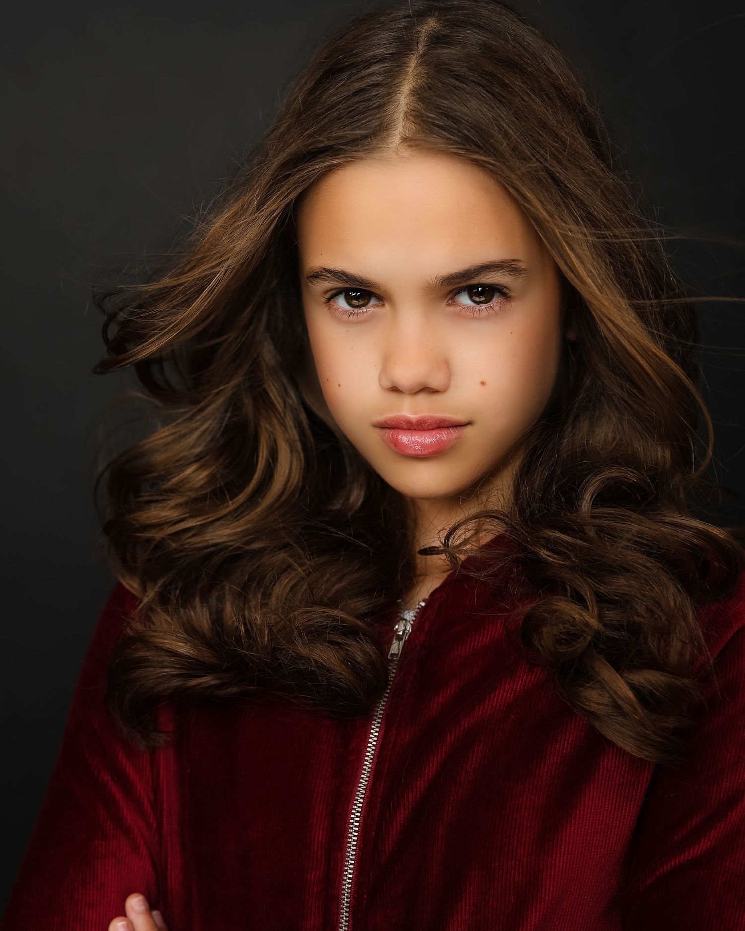 Professional portrait in red velvet jacket against dark background with styled brown waves.