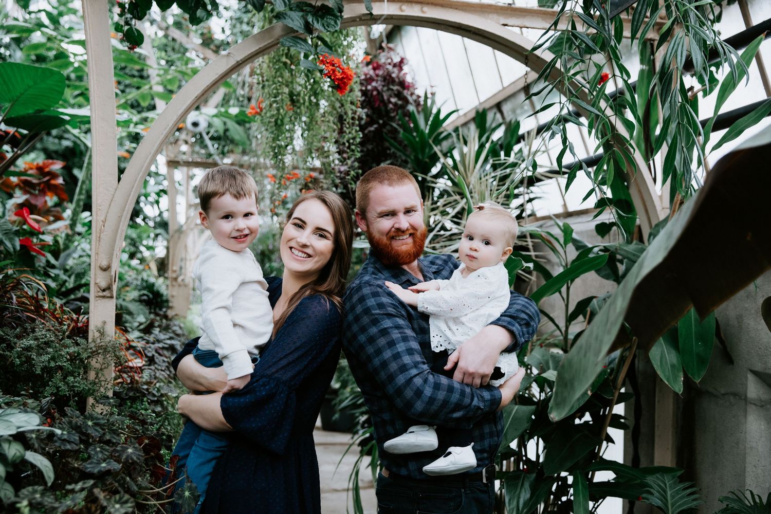 Family portrait in a lush greenhouse with tropical plants and hanging vines.