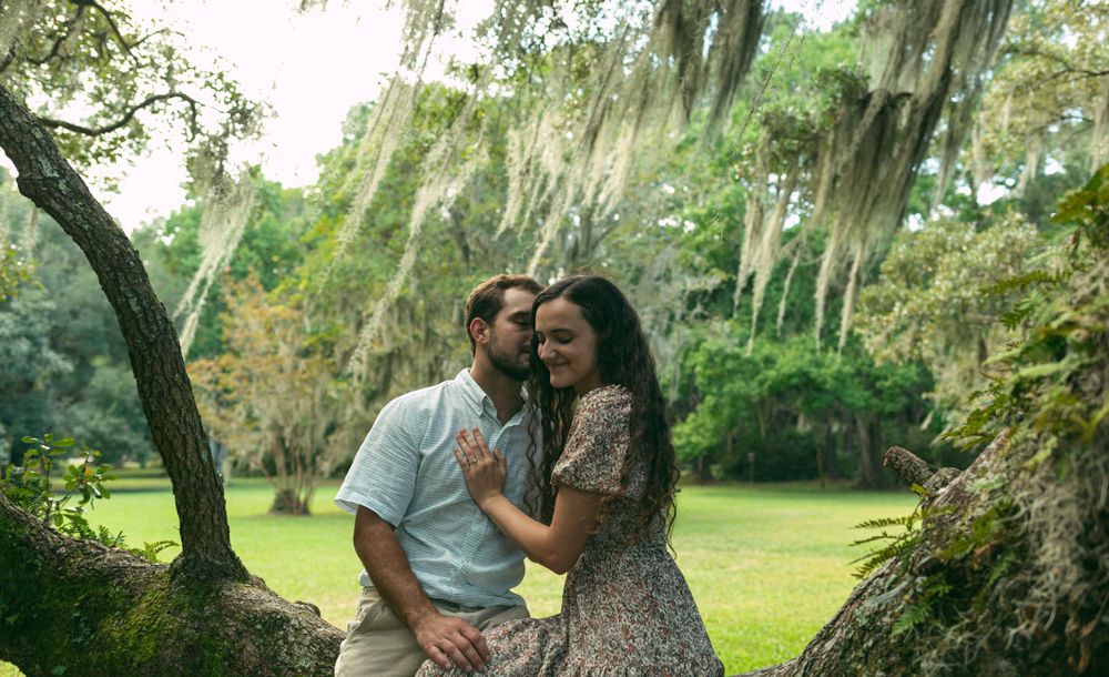A couple shares laughter while perched on a moss-draped tree in a sunlit garden.