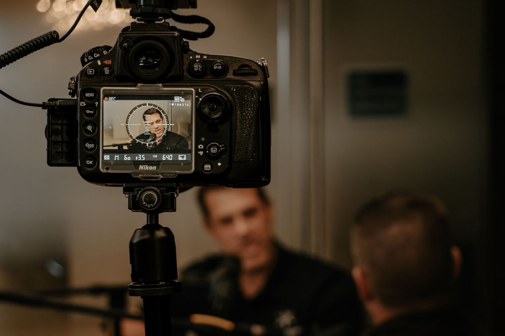 Digital camera mounted on tripod recording interview with people sitting in dimly lit room during production shoot.