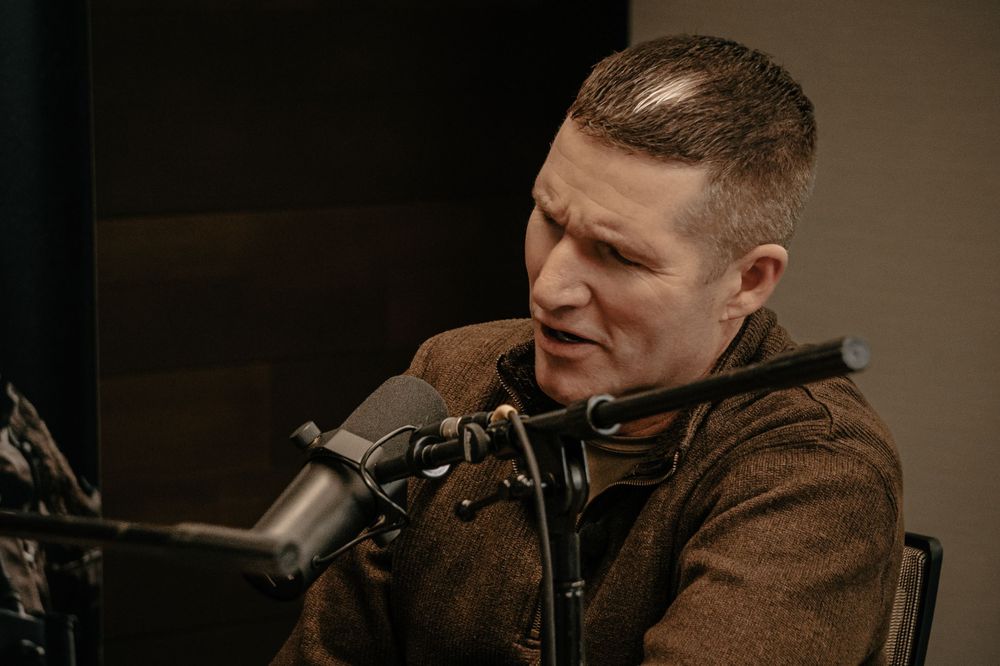 Someone speaks into a microphone during a podcast recording session while wearing a brown jacket in dim lighting.