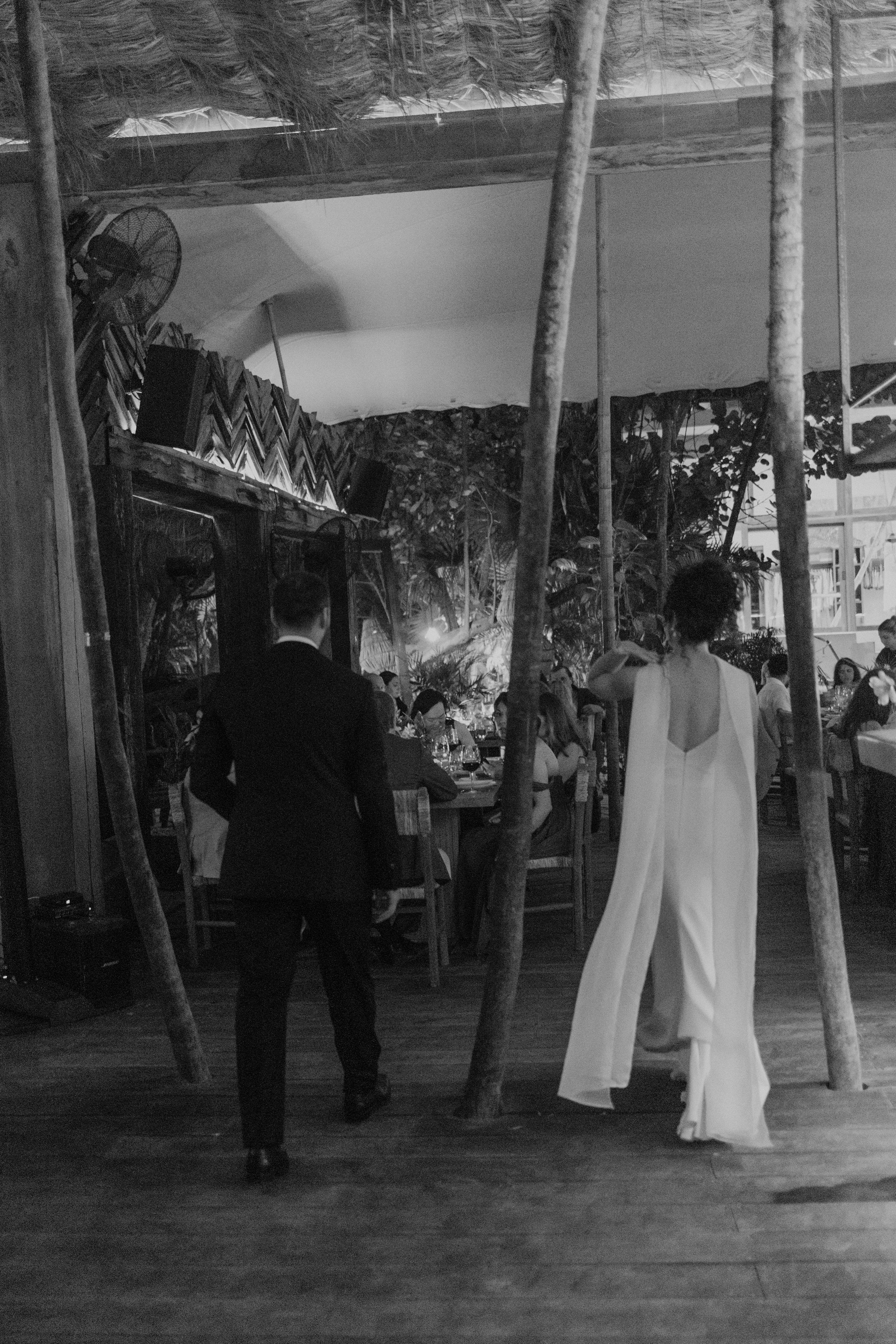 Black and white photo of a bride and groom walking into their wedding reception venue surrounded by tropical trees.