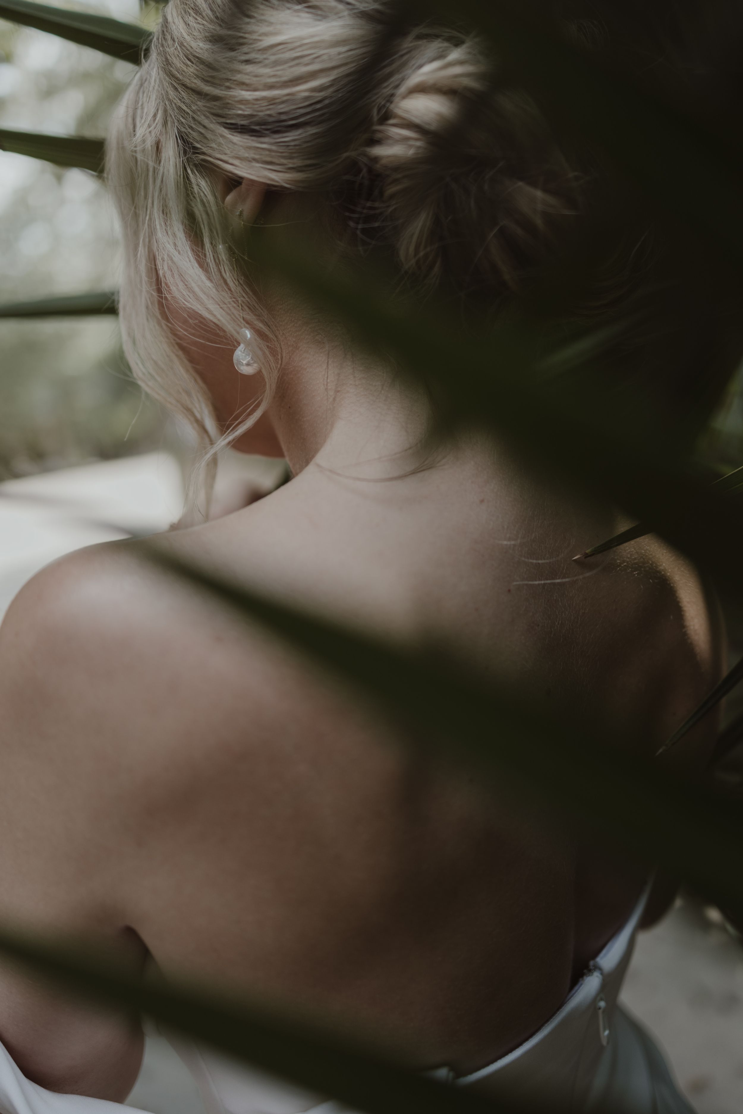 Bride with elegant updo and pearl earring, backless wedding dress, photographed through green foliage.