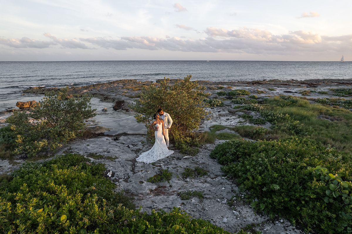 Aerial view of a bride and groom embracing on a rocky oceanside shore surrounded by lush greenery at sunset.