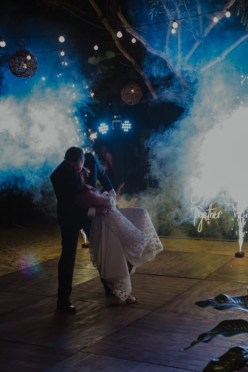 Couple sharing first dance on wooden floor surrounded by blue smoke and string lights at an outdoor wedding reception.