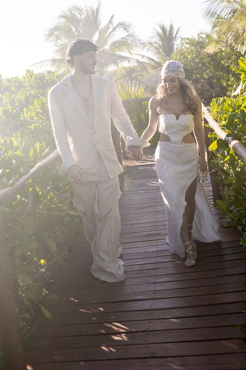 A couple in white wedding attire walks hand-in-hand on a tropical wooden boardwalk surrounded by lush greenery.