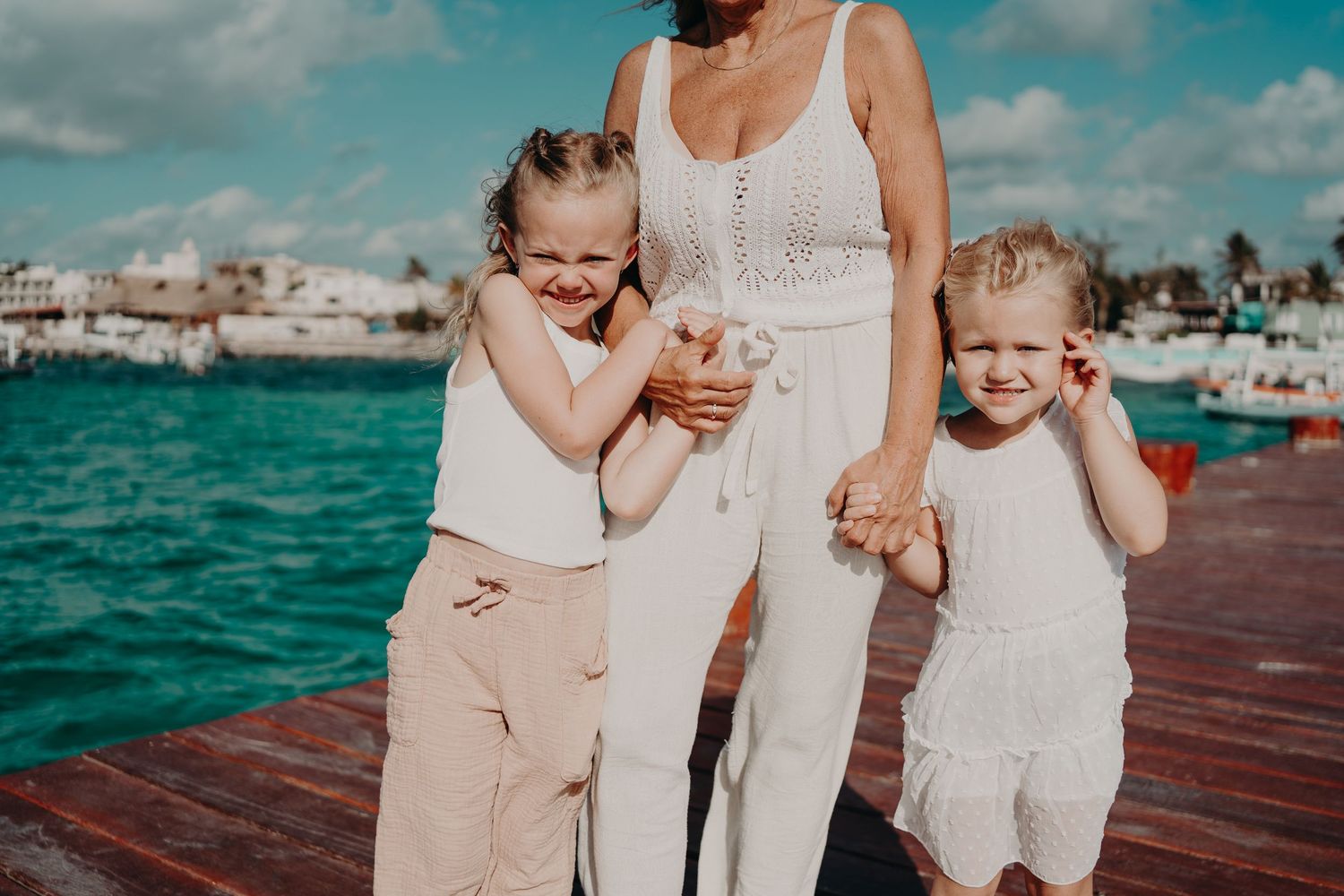 Two children in light colored outfits share a joyful moment with an adult on a wooden dock. Family photography in Puerto Morelos.