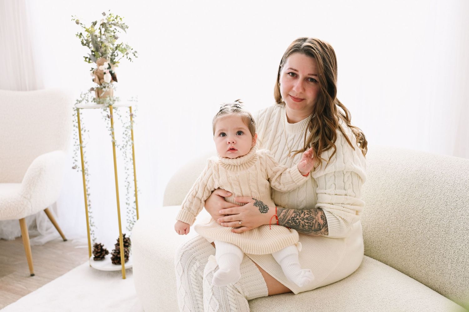 Mother and child wearing cream sweaters share tender moments on a white couch in a bright minimalist setting.