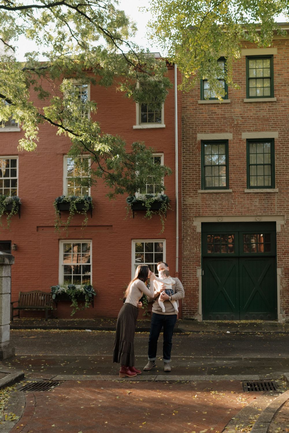 People gather outside a historic red brick building with black doors and windows on a tree-lined cobblestone street.