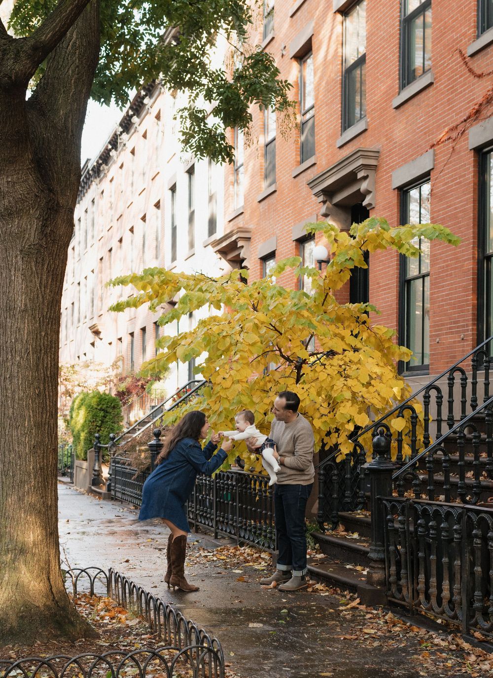 People gather on a historic brownstone-lined street in autumn with fallen leaves scattered on the sidewalk.