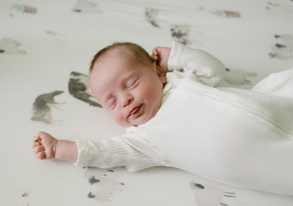 A baby in white sleepwear lies on a light-colored surface with scattered feathers around them.