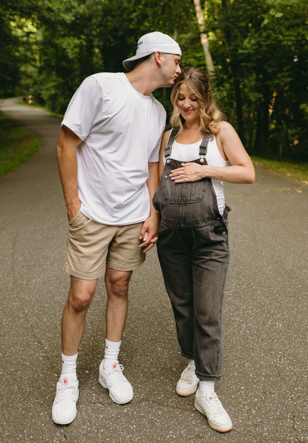 A couple walking on a tree-lined path, with one wearing a white t-shirt and shorts and the other in denim overalls.