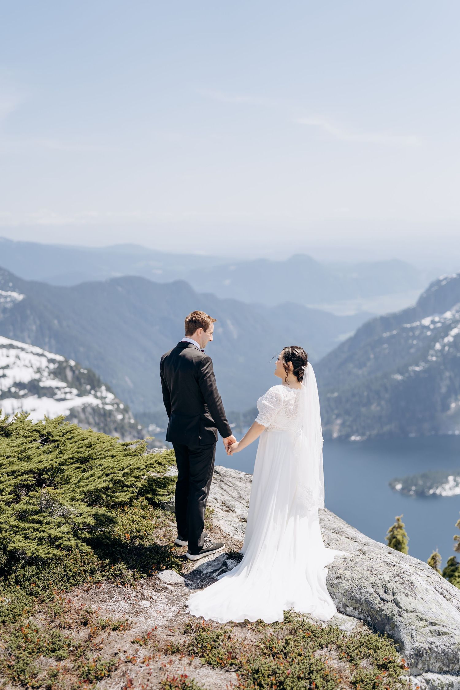 Newlyweds stand on mountain peak overlooking scenic lake and mountain ranges on their wedding day.