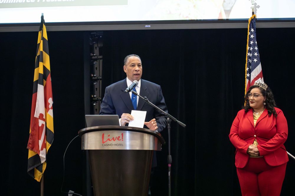 A series of speakers address an audience from behind podiums at a formal event with American and Maryland state flags visible.